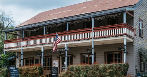 A two-story building with a red roof, American flag, and outdoor seating, surrounded by greenery and decorative lights.