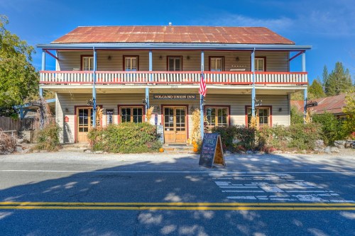 A charming two-story inn with a rustic facade, surrounded by greenery and a clear blue sky. An American flag is displayed.