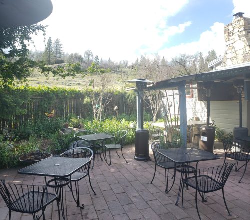 A serene outdoor patio with metal tables and chairs, surrounded by greenery and a scenic hillside in the background.
