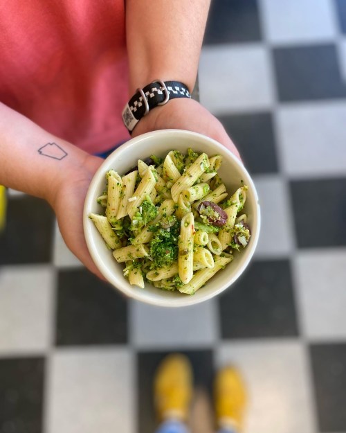 A person holds a bowl of penne pasta with pesto and vegetables, set against a checkered floor.