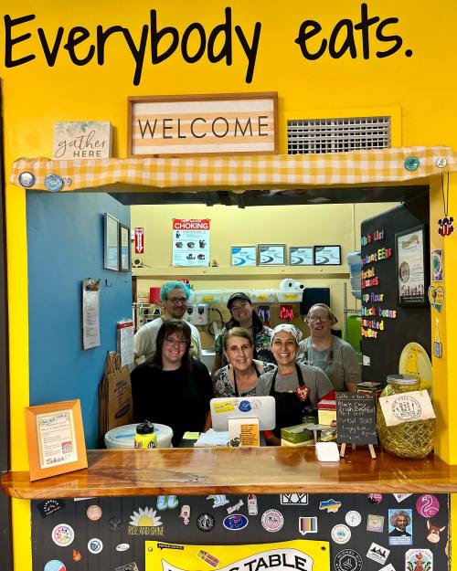 A welcoming kitchen scene with staff smiling behind a counter, surrounded by colorful decor and a "Welcome" sign.