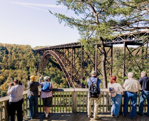 Visitors stand on a wooden overlook, admiring a large bridge spanning a lush, green valley under a clear blue sky.