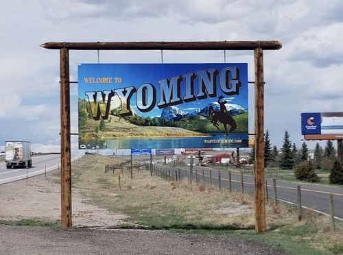 Sign welcoming visitors to Wyoming, featuring mountains and a cowboy silhouette, with highway and trucks in the background.
