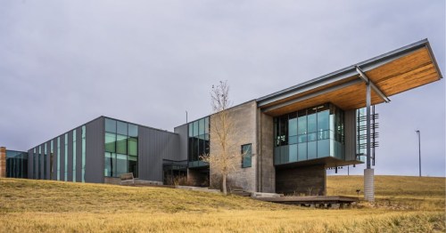 Modern building with large glass windows and a wooden overhang, set against a cloudy sky and grassy landscape.