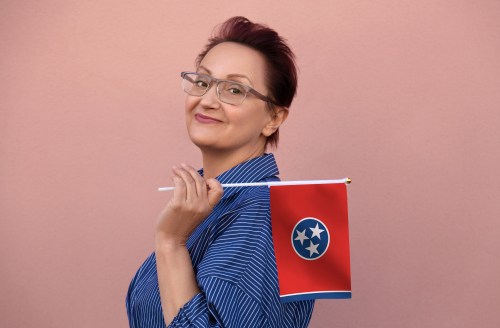 Portrait of a middle-aged woman (40–50) standing on a street with a state flag over a pink wall in the background.