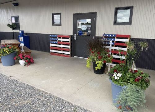 Entrance to a building featuring wooden American flag decor and colorful flower pots on a concrete walkway.