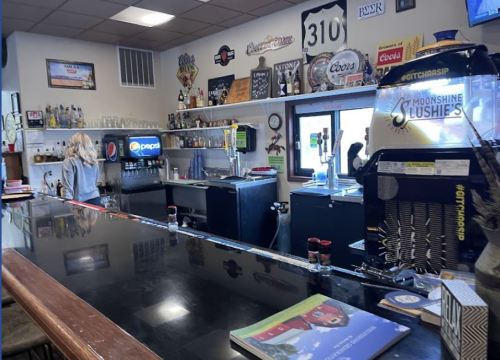A bar counter with drinks, a bartender, and various signs and decorations on the walls.