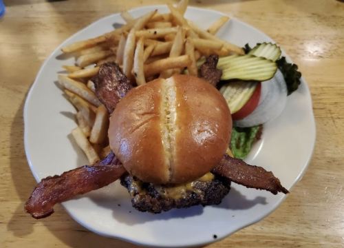 A burger with bacon on top, served with fries, lettuce, tomato, pickles, and onion on a white plate.