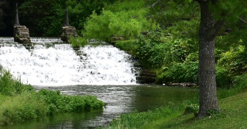 A serene waterfall cascades into a calm river, surrounded by lush greenery and trees.