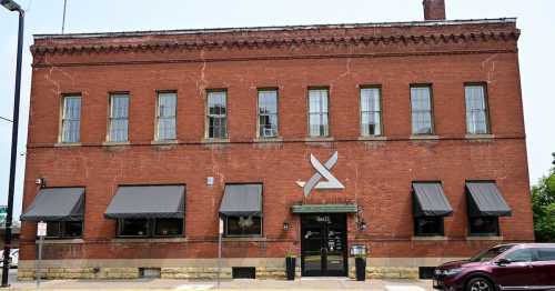 A red brick building with large windows and black awnings, featuring a modern logo above the entrance.