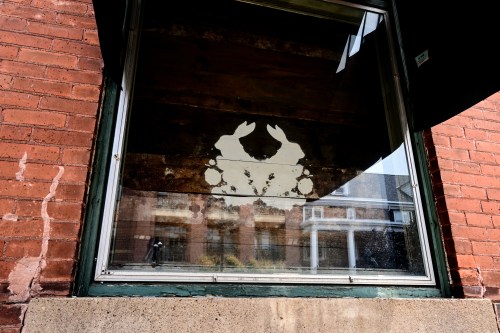 A window with a faded, artistic crab silhouette against a brick wall, reflecting the surrounding buildings.