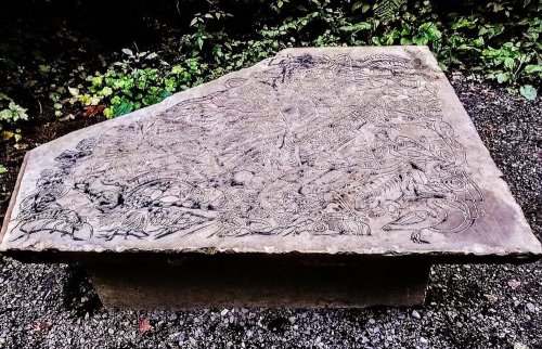 A large stone slab with intricate carvings, surrounded by greenery and gravel.