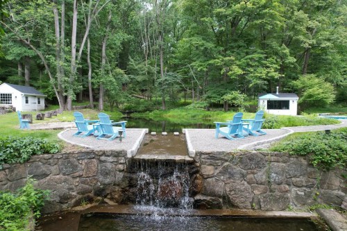 A serene outdoor scene featuring blue chairs by a pond, surrounded by lush greenery and a small waterfall.