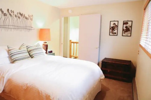 Cozy bedroom with a white bed, decorative pillows, a lamp, and artwork on the walls. A door leads to another room.