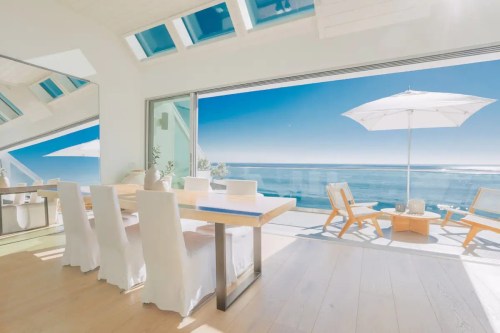 Bright, modern dining area with a large table, white chairs, and ocean views through expansive glass doors.