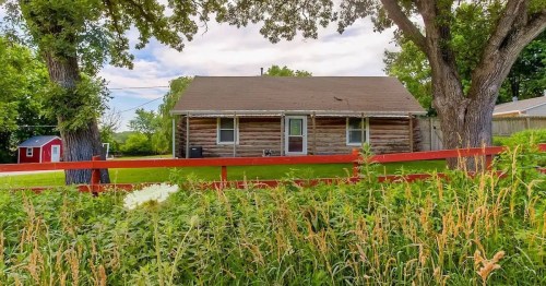 A rustic log cabin surrounded by greenery and a red fence, with trees and a small shed in the background.