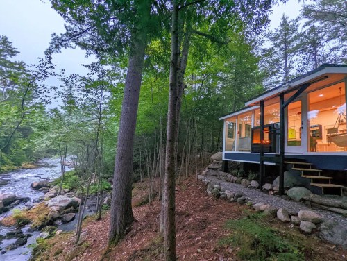 A cozy cabin by a river, surrounded by trees, with large windows and a stone pathway leading to the entrance.