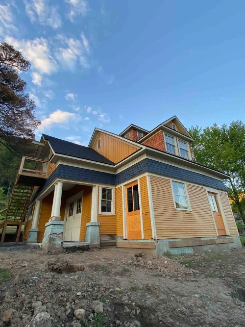 A partially constructed house with orange siding, white trim, and a blue roof, set against a clear sky and trees.