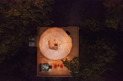 Aerial view of a geodesic dome cabin surrounded by trees, featuring a wooden deck and outdoor seating.