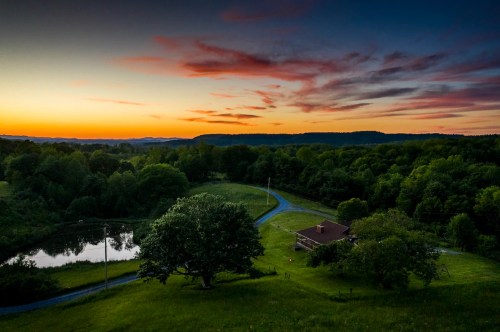 A serene landscape at sunset, featuring a house, pond, and lush greenery under a colorful sky.