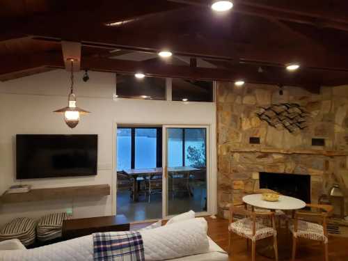 Cozy living room with a stone fireplace, TV, and a view of a lake through large windows. Modern decor and seating.