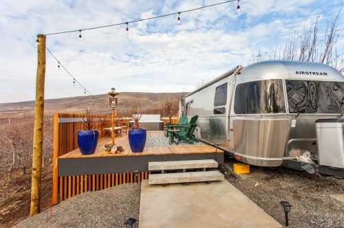 A silver Airstream trailer beside a wooden deck with chairs, planters, and string lights against a scenic landscape.