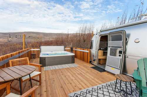 A cozy outdoor deck featuring a hot tub, wooden furniture, and an Airstream trailer against a scenic landscape.