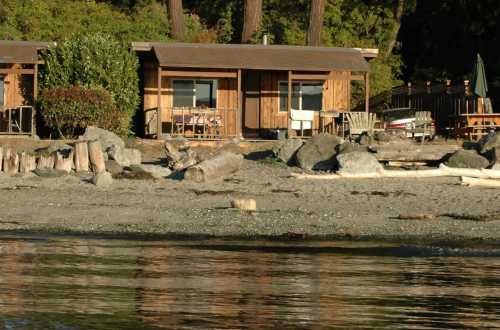 A wooden cabin by the beach, surrounded by trees and rocks, with a calm water reflection in the foreground.