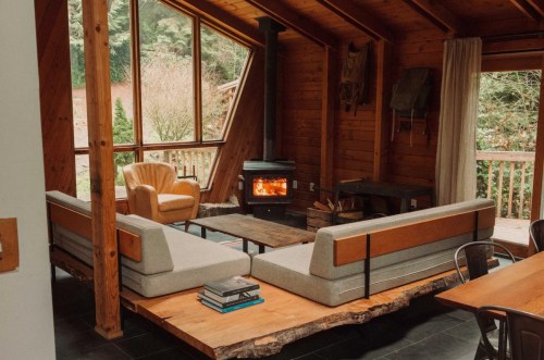 Cozy wooden cabin interior with a modern sofa, armchair, and a wood stove, surrounded by large windows.