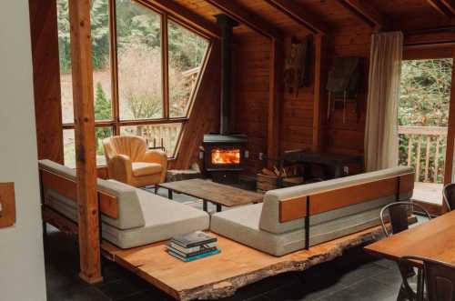 Cozy wooden cabin interior with a modern sofa, armchair, and a wood stove, surrounded by large windows.