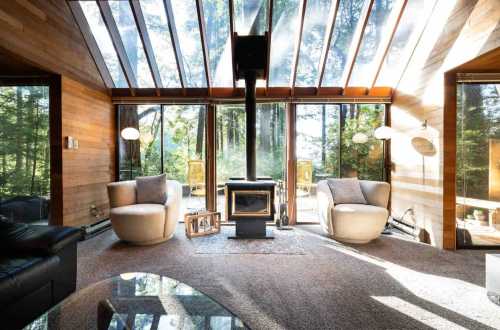 Cozy living room with large windows, two modern chairs, a wood stove, and a view of a forested area outside.