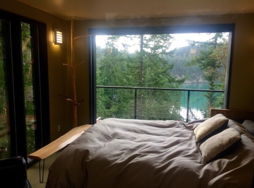 Cozy bedroom with a large window overlooking a serene lake and lush trees. Natural light fills the space.