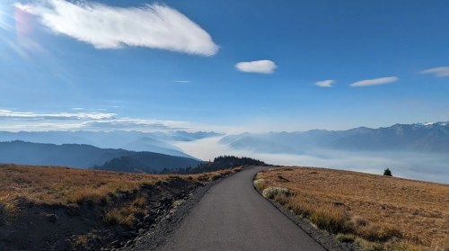A winding road leads through a mountainous landscape under a clear blue sky, with distant peaks shrouded in mist.
