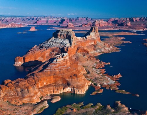 Aerial view of colorful rock formations and blue water at Lake Powell, surrounded by rugged cliffs and canyons.