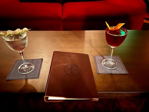 Two cocktails on a table with a menu, set against a backdrop of a red couch. One drink is clear, the other is dark red.