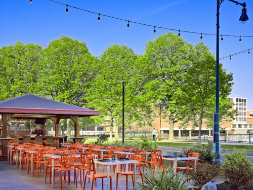 Outdoor dining area with orange chairs, tables, and string lights, surrounded by lush green trees and a clear blue sky.