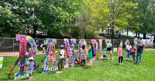 A colorful "BEACH" sign with children and adults painting it in a park, surrounded by greenery and trees.