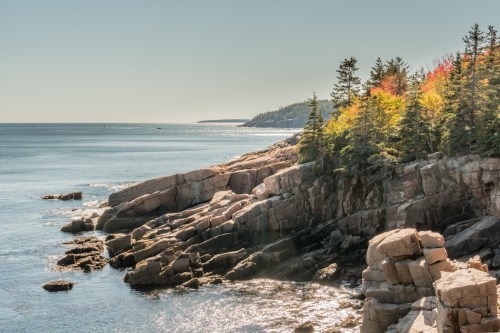 Coastal view with rocky cliffs, calm waters, and trees displaying autumn colors under a clear sky.