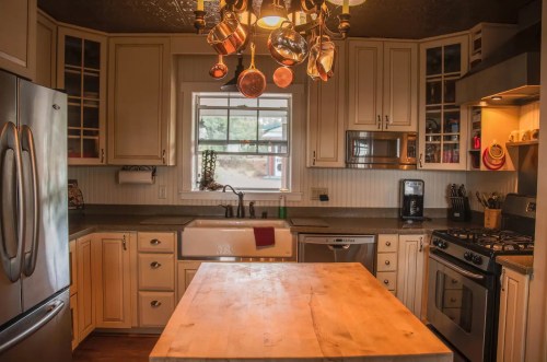 A cozy kitchen featuring a wooden island, stainless steel appliances, and hanging pots above a sink by a window.