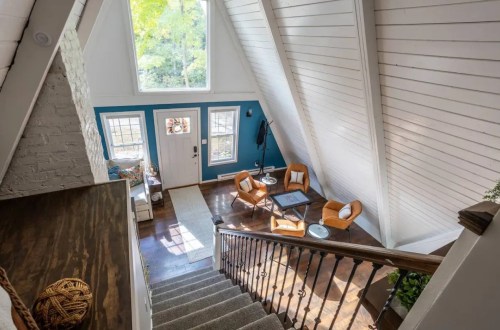 A cozy, modern living room with high ceilings, large windows, and stylish orange chairs, viewed from a staircase.
