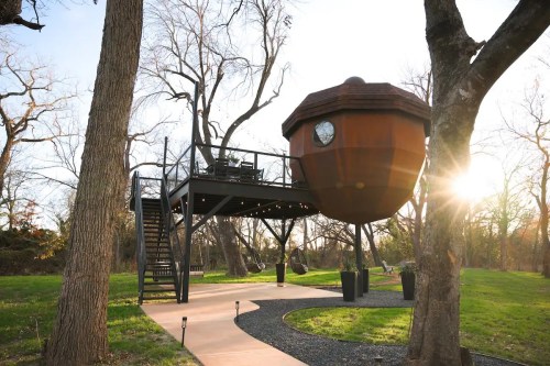 A unique treehouse shaped like a large, round structure, elevated on stilts, surrounded by trees and a pathway.