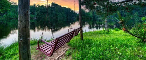 A red swing hangs by a calm lake, surrounded by lush greenery and trees, under a colorful sunset sky.