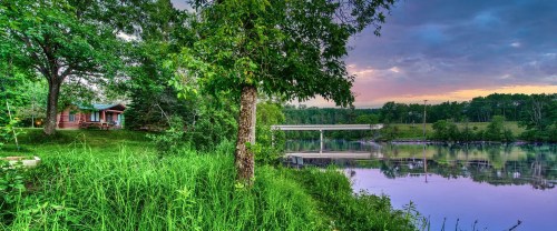 A serene lakeside scene with a log cabin, lush greenery, and a calm water surface reflecting a colorful sky.