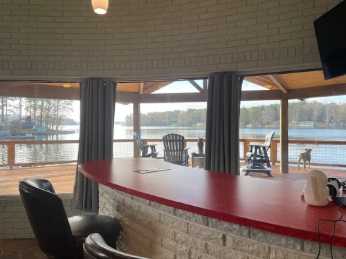 Cozy interior with a red countertop, overlooking a lake through large windows and a wooden deck with chairs.