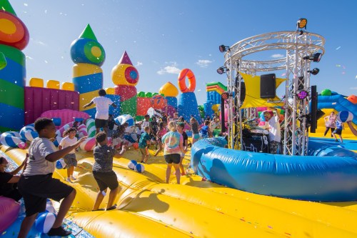 A vibrant inflatable playground filled with children playing, balloons, and a DJ setup under a clear blue sky.