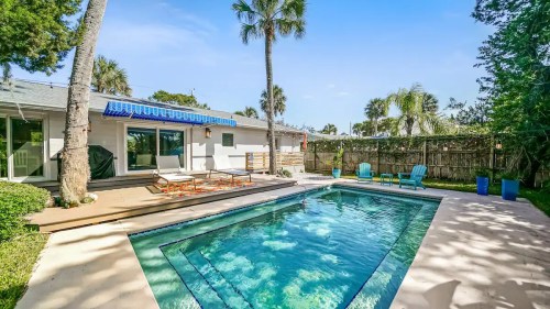 A sunny backyard with a pool, lounge chairs, and palm trees, surrounded by greenery and a blue sky.