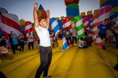 A joyful person celebrates on a colorful inflatable bounce house, surrounded by a crowd of excited people.