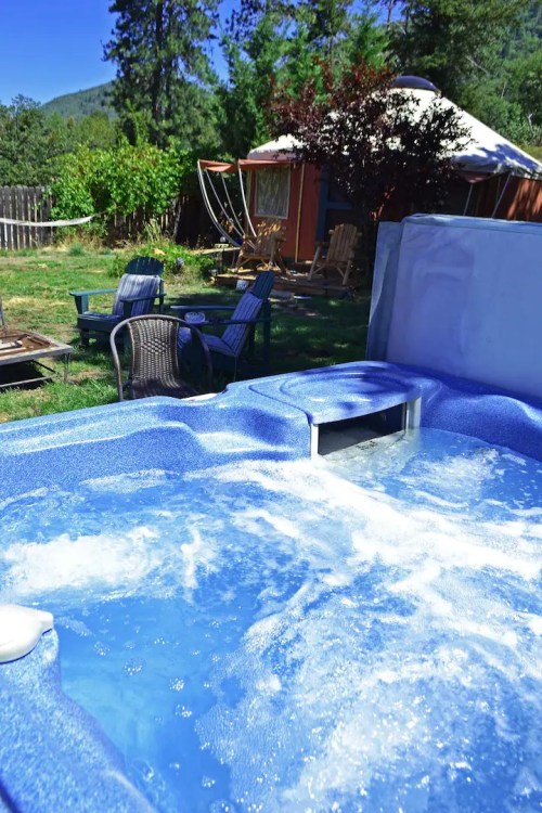 A bubbling hot tub in a sunny backyard with chairs and a cabin in the background, surrounded by trees.