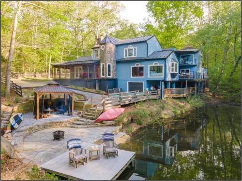 A blue house by a pond, surrounded by trees, with a patio, seating area, and a kayak on the shore.