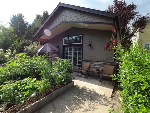 A cozy house with a garden, featuring a patio with chairs and an umbrella, surrounded by lush greenery.